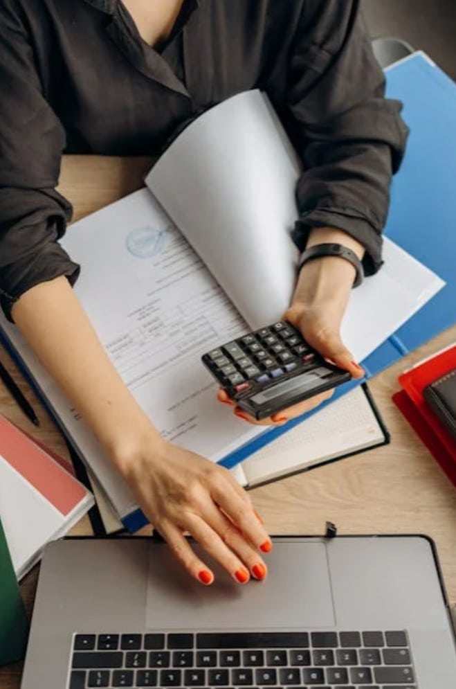 Multitasking at Work: Balancing Paperwork and Technology A woman in a black shirt using a calculator and laptop while working with paperwork and folders spread across a desk.